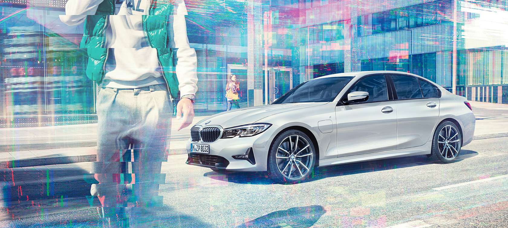 BMW 3 Series Sedan, side shot BMW 330e. Three-quarter side shot of the standing BMW 330e Sedan in front of an urban glass building with walking passengers.