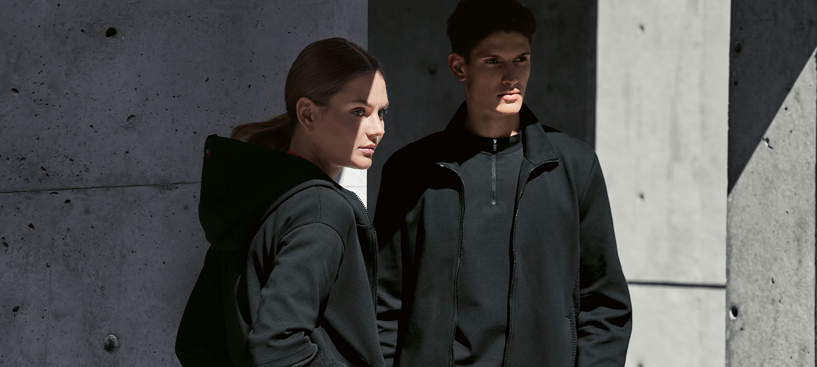 BMW M Sweat Jacket for Ladies and BMW M Sweat Jacket for Men. A couple is standing in the shadow. A woman is presenting the BMW M Sweat Jacket for ladies and a man is wearing the BMW M Sweat Jacket for men.